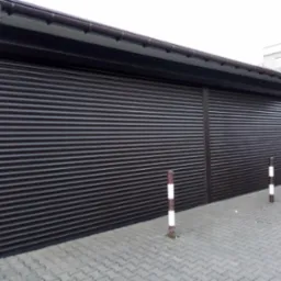 Zewnętrzne, brązowe rolety zabezpieczające na budynku z ciemnym orynnowaniem, widoczne z poziomu chodnika wyłożonego kostką.