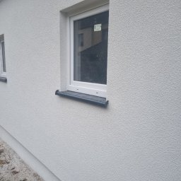 Kw-Kris Bud - Świeżo otynkowana, jasna elewacja budynku z dwoma oknami o białych ramach i grafitowych parapetach. Na szybie jednego z okien widoczna naklejka z informacjami. W tle odbicie budynku.