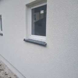 Świeżo otynkowana, jasna elewacja budynku z dwoma oknami o białych ramach i grafitowych parapetach. Na szybie jednego z okien widoczna naklejka z informacjami. W tle odbicie budynku.