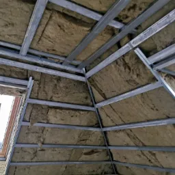 Montaż izolacji z wełny mineralnej w poddaszu, widoczna metalowa konstrukcja szkieletowa i okno dachowe.