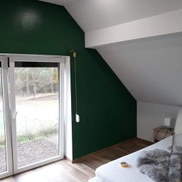 Kw-Kris Bud - Pokój na poddaszu po remoncie: ściana pomalowana na ciemnozielono, drewnopodobne panele na podłodze, białe okno balkonowe, widok na zewnątrz.