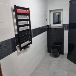 Nowoczesna łazienka z biało-czarnymi płytkami, szarymi płytkami podłogowymi i czarną armaturą: toaleta i grzejnik drabinkowy z czerwonym akcentem na górze.