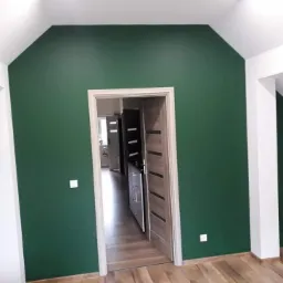 Pomieszczenie po malowaniu ścian na ciemnozielony kolor, widoczne białe wykończenia sufitu i drzwi, podłoga drewniana, widok na wnętrze przez otwarte drzwi.