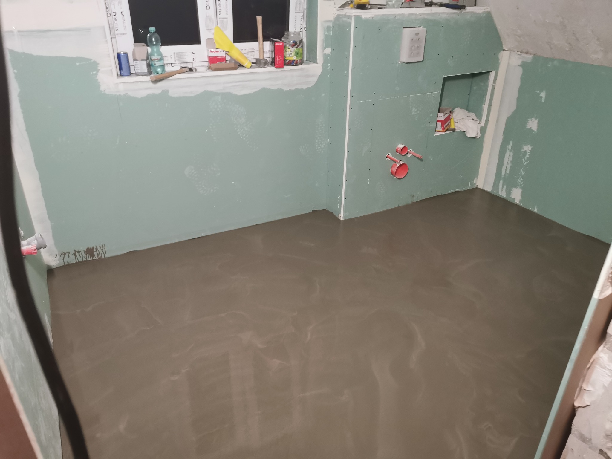 Wyrównana, wylewana podłoga w łazience z panelami kartonowo-gipsowymi na ścianach, widoczne instalacje wodne i narzędzia na parapecie okna.