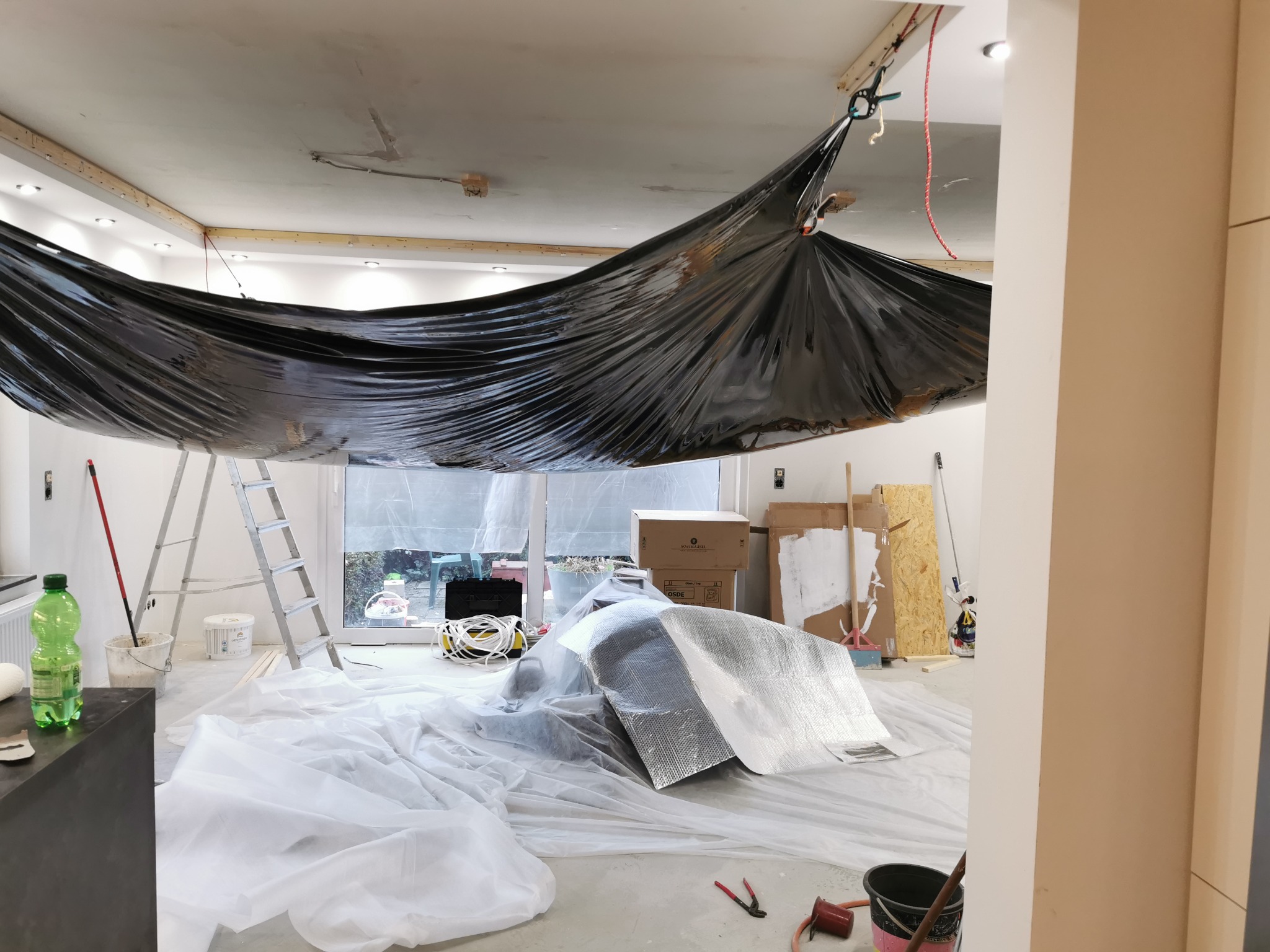 Przygotowanie pomieszczenia do prac wykończeniowych: folia ochronna zawieszona pod sufitem, zabezpieczona podłoga, drabina, narzędzia malarskie w tle.