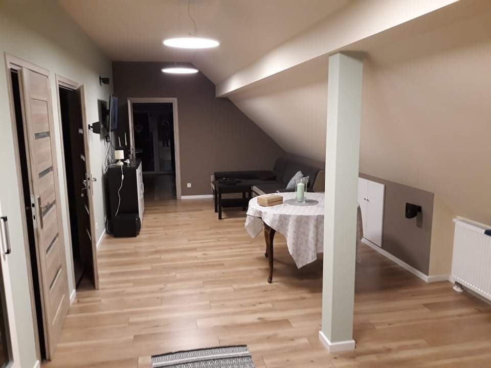 Wykończone poddasze ze skośnym sufitem, drewnianą podłogą, jasnymi ścianami, okrągłym stołem przy filarze i widocznym fragmentem sofy w tle.