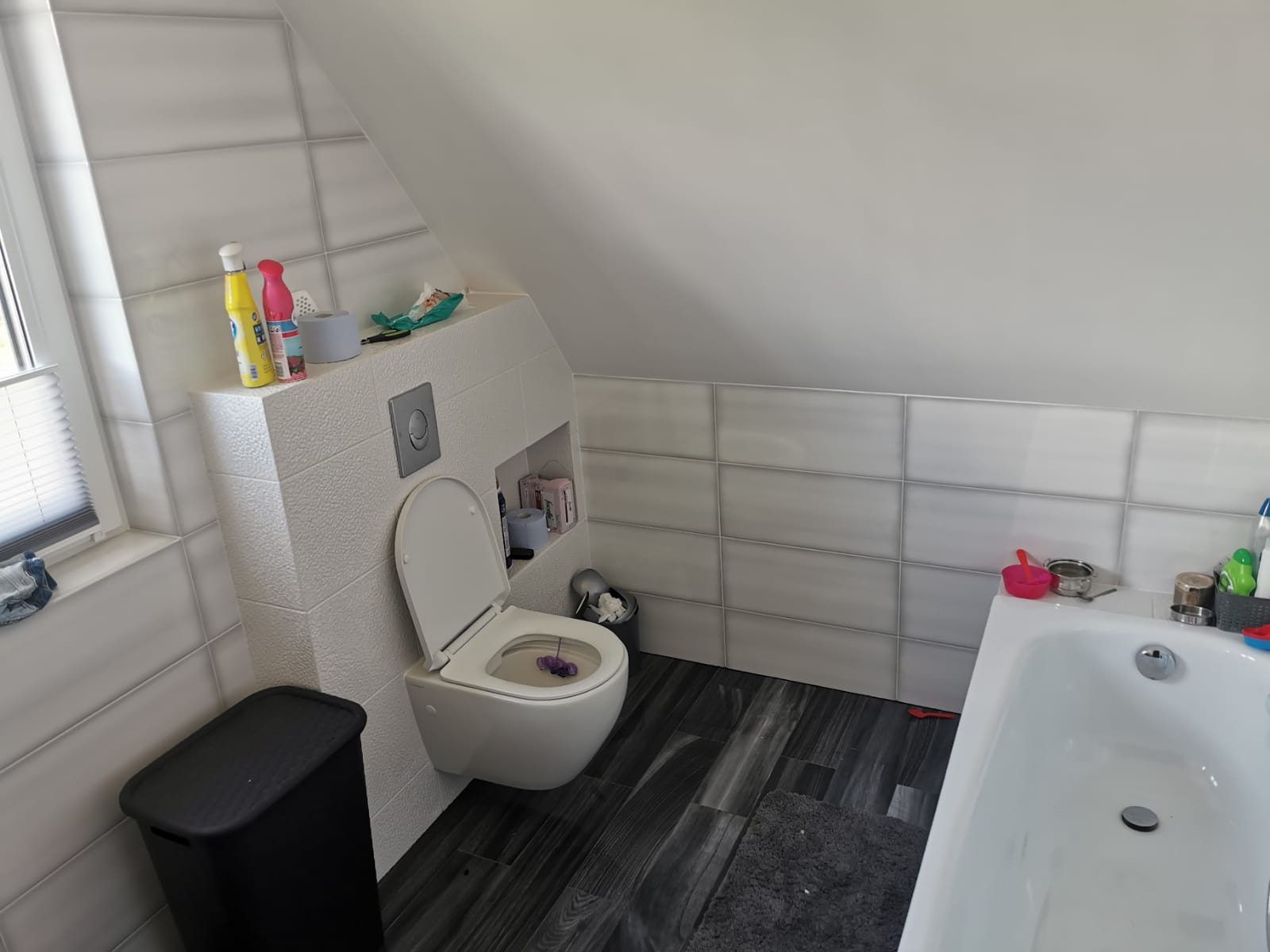 Biała toaleta podwieszana z otwartą klapą w łazience na poddaszu, obudowana płytkami o fakturze baranka, z szarą podłogą imitującą drewno i widocznymi akcesoriami łazienkowymi.