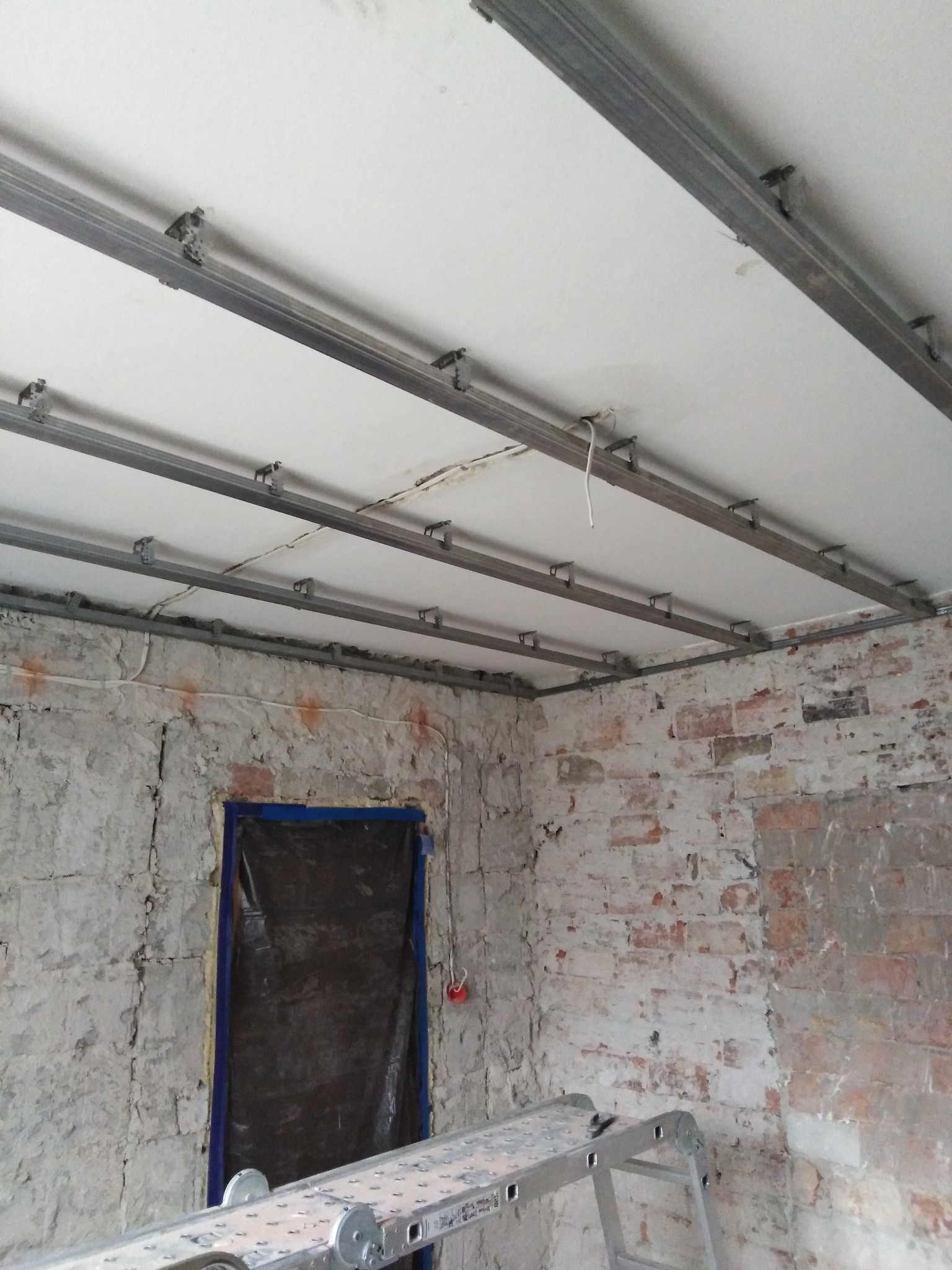 Montaż stelaża pod płytę gipsowo-kartonową na suficie w surowym pomieszczeniu z widoczną drabiną i zakrytymi drzwiami.