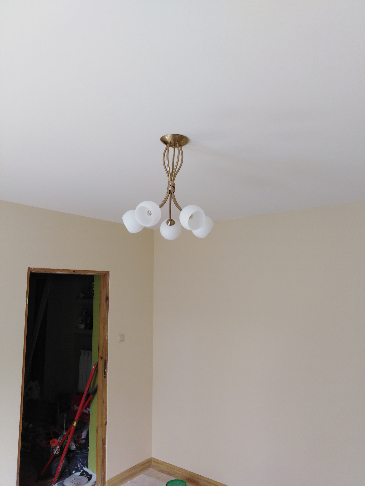 Świeżo pomalowane ściany w odcieniach beżu z zamontowanym żyrandolem z białymi kloszami i złotymi ramionami, widoczny fragment wykończonej podłogi i otwarte drzwi do zagraconego pomieszczenia.