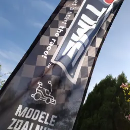 Flaga z szachownicą i napisem 'Modele Zdalnie Sterowane' oraz logo firmy na tle błękitnego nieba i zieleni.