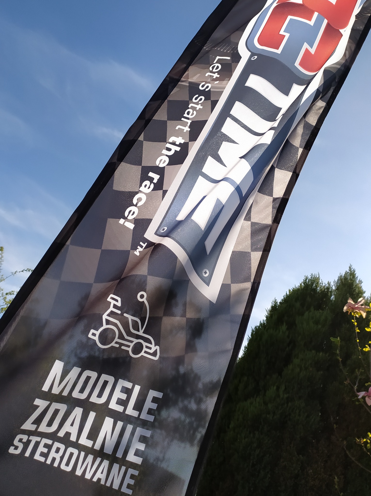 Flaga z szachownicą i napisem 'Modele Zdalnie Sterowane' oraz logo firmy na tle błękitnego nieba i zieleni.