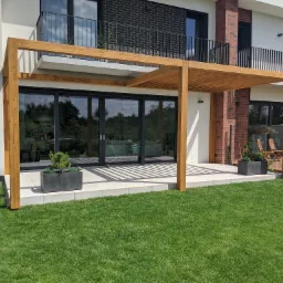 Drewniana konstrukcja zadaszenia tarasu przy nowoczesnym domu z przesuwnymi drzwiami, z roślinami w donicach i zadbanym trawnikiem.