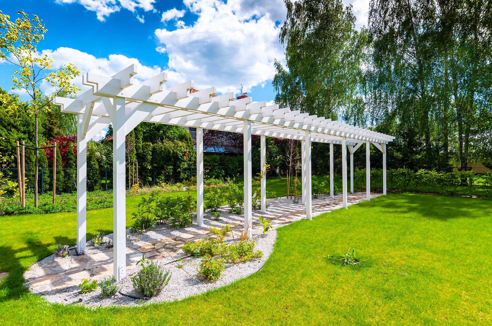 Długa, biała konstrukcja z belek tworząca zadaszenie nad ścieżką z jasnych kamieni w zadbanym ogrodzie, z widokiem na drzewa i błękitne niebo z chmurami.