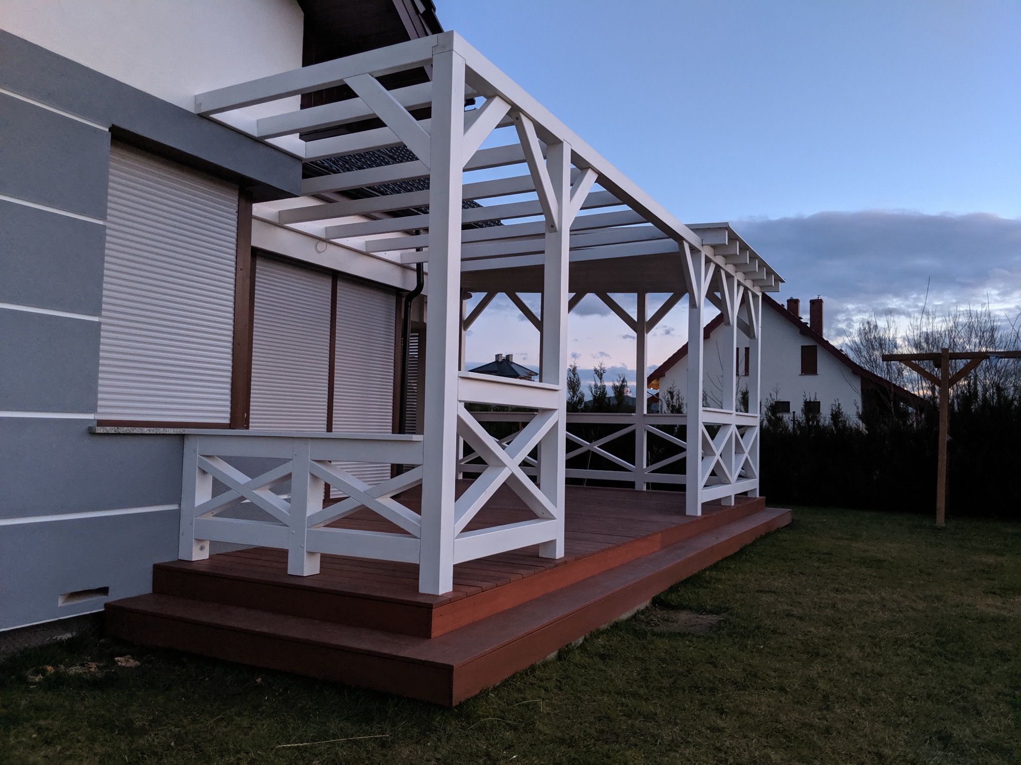 Biała konstrukcja z drewnianym podestem przy szarym domu, widok na ogród i zachodzące słońce.