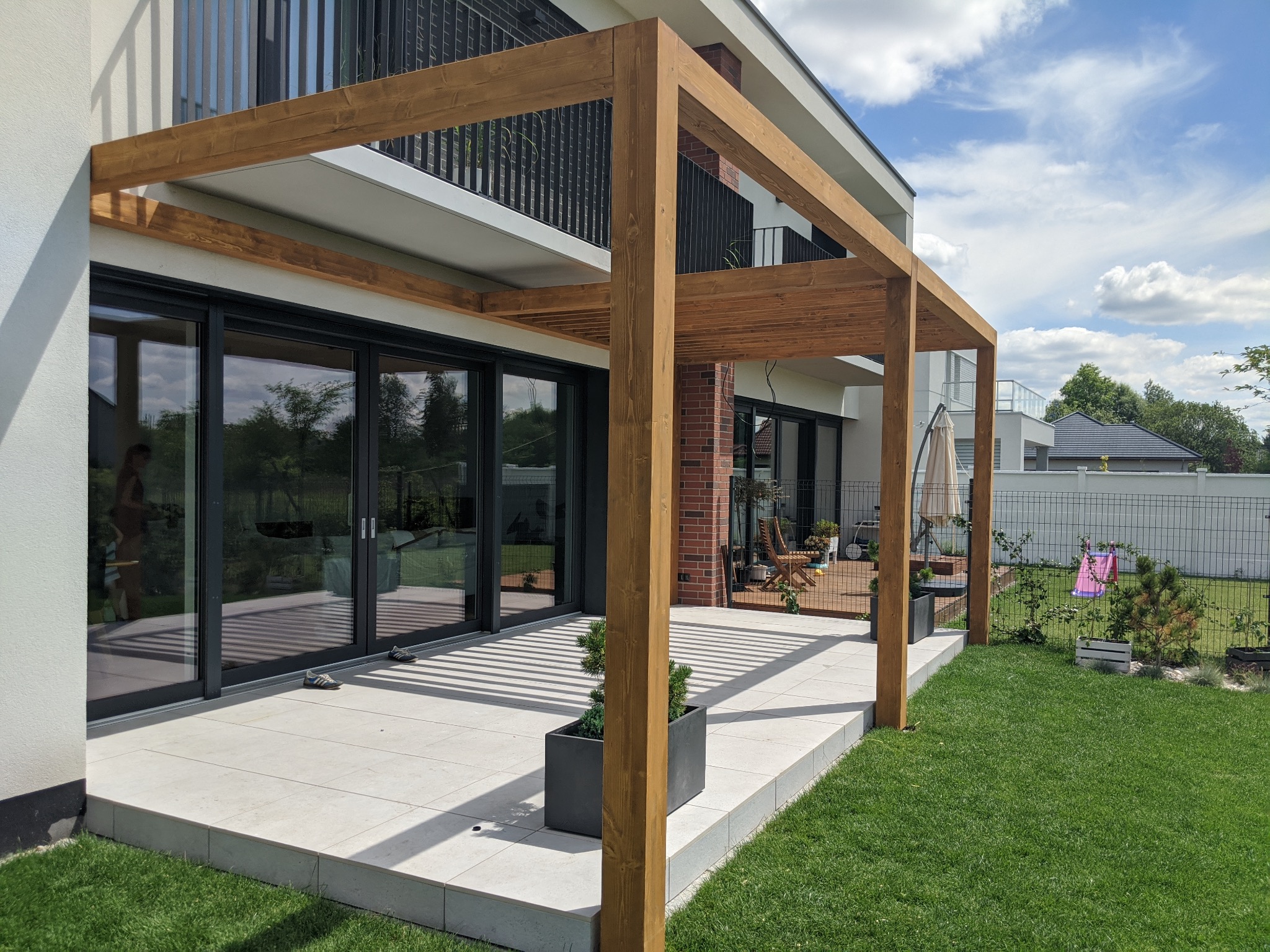 Nowoczesna, drewniana konstrukcja zadaszenia tarasu, z widocznymi słupami i belkami, tworząca cień na jasnej, kamiennej posadzce tarasu przylegającego do budynku z dużymi przesuwnymi oknami.