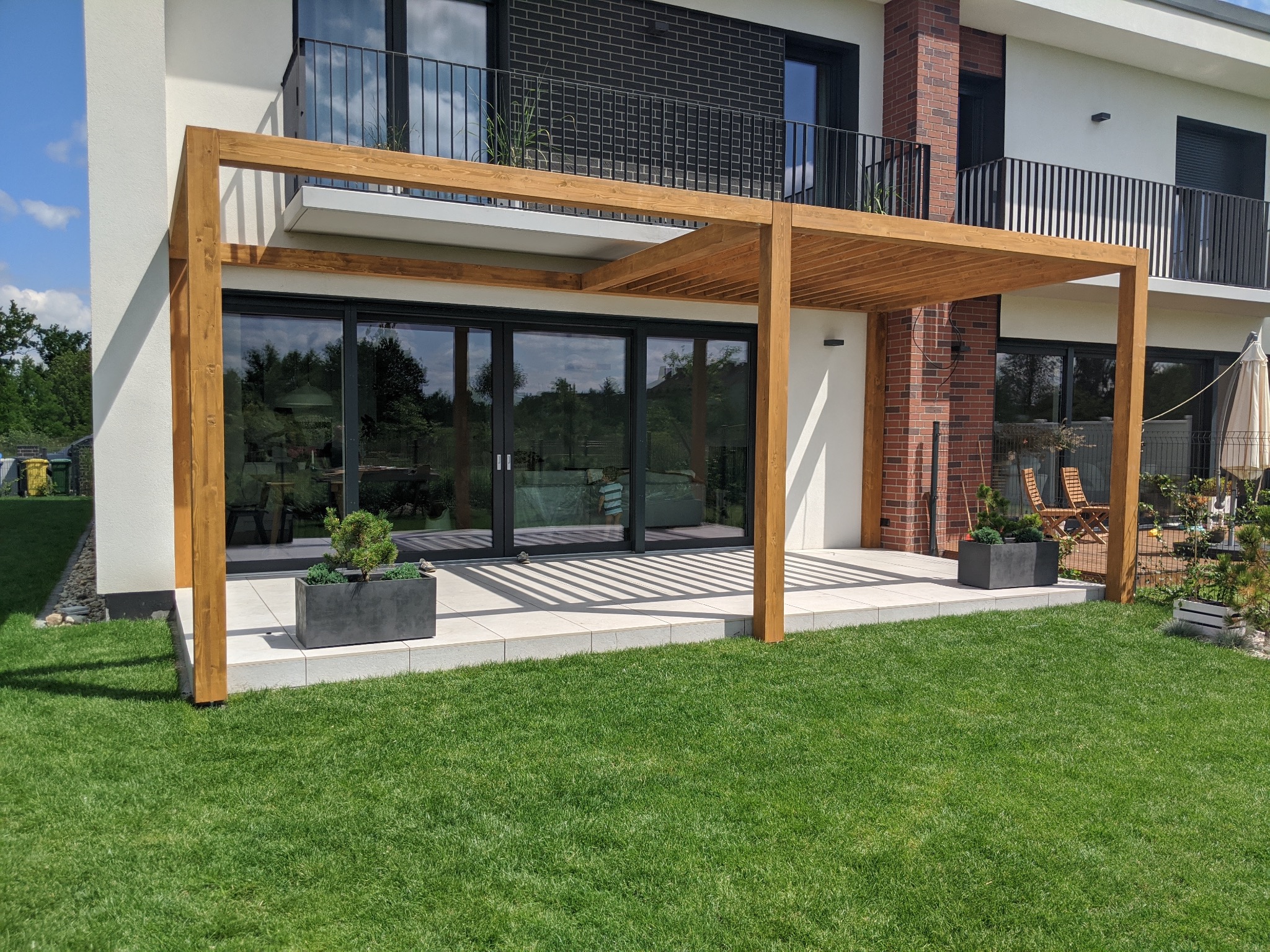 Drewniana konstrukcja zadaszenia tarasu przy nowoczesnym domu z przesuwnymi drzwiami, z roślinami w donicach i zadbanym trawnikiem.