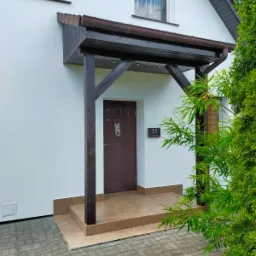 Wejście do domu po remoncie: ocieplona biała elewacja, brązowy drewniany daszek z kolumnami, płytki na schodach i chodniku, numer domu '11' przy drzwiach.