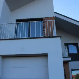 Nowoczesny dom jednorodzinny z białymi ścianami, balkonem z metalową balustradą, garażem i fragmentem elewacji wykończonym drewnem.
