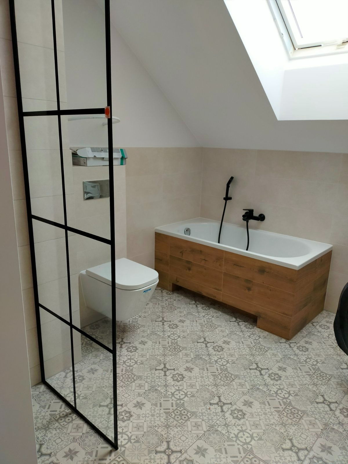 Nowoczesna łazienka na poddaszu z kabiną prysznicową typu walk-in z czarnymi profilami, wiszącą toaletą, wanną obudowaną drewnem i wzorzystą, szarą podłogą.