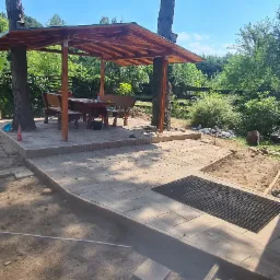 Drewniana altana z meblami ogrodowymi, wbudowana w dwa drzewa, na podwyższonym tarasie z kostki brukowej, w tle zieleń ogrodu.