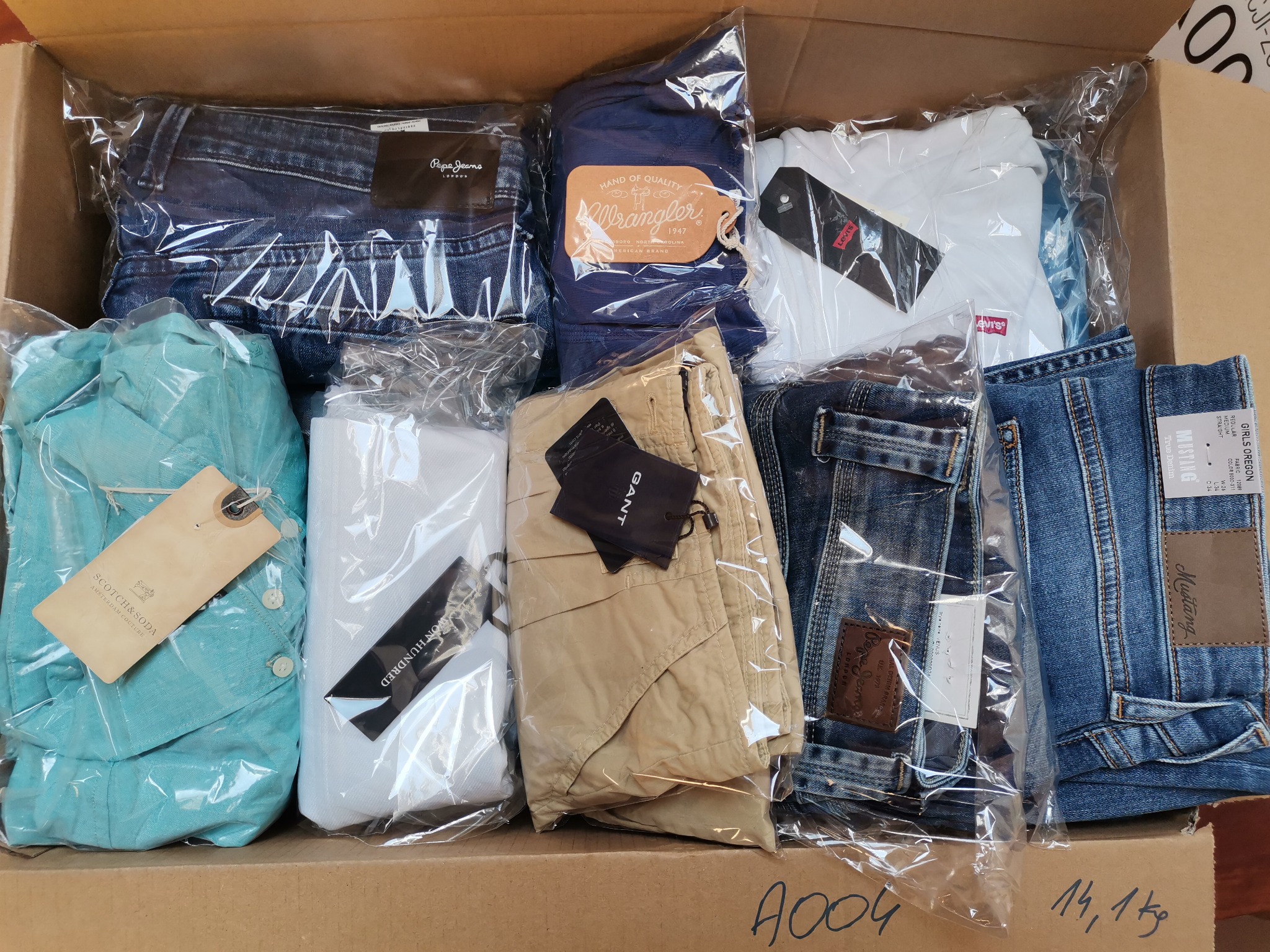 Kartonowe pudło wypełnione różnorodną odzieżą markową, każda sztuka zapakowana w przezroczysty plastik, widoczne dżinsy Pepe Jeans, Wrangler, Levi's, koszula Scotch & Soda, spodnie Gant i inne,...