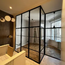 Nowoczesna, przeszklona ścianka działowa z czarnymi szprosami, oddzielająca przestrzeń biurową z parkietem w jodełkę i designerskim oświetleniem. Widoczny fragment biurka i krzesła w strefie...