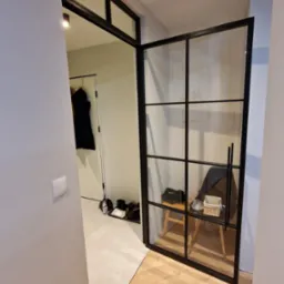 Nowoczesne, przeszklone drzwi wewnętrzne z czarnymi szprosami, oddzielające przedpokój z wieszakiem na ubrania i płytkami podłogowymi od pokoju z drewnianą podłogą ułożoną w jodełkę.