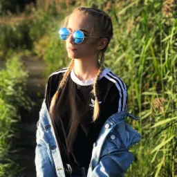 Portret młodej kobiety w okularach przeciwsłonecznych z odblaskami nieba i trzcin, ubrana w czarny t-shirt z białymi paskami i jeansową kurtkę, stojącej na tle wysokich traw.