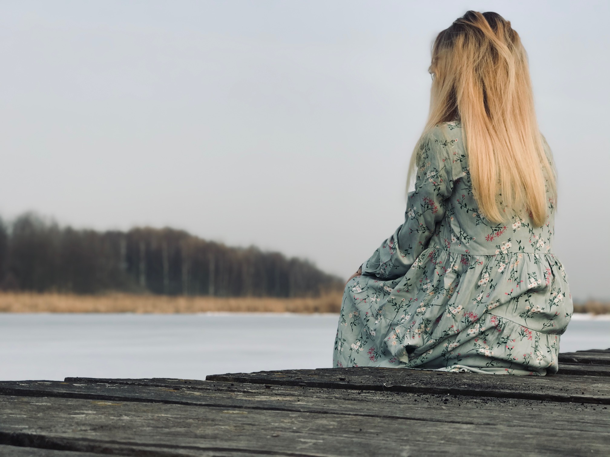 Kobieta w kwiecistej sukience siedzi tyłem na drewnianym pomoście nad zamarzniętym jeziorem, w tle widoczny las.