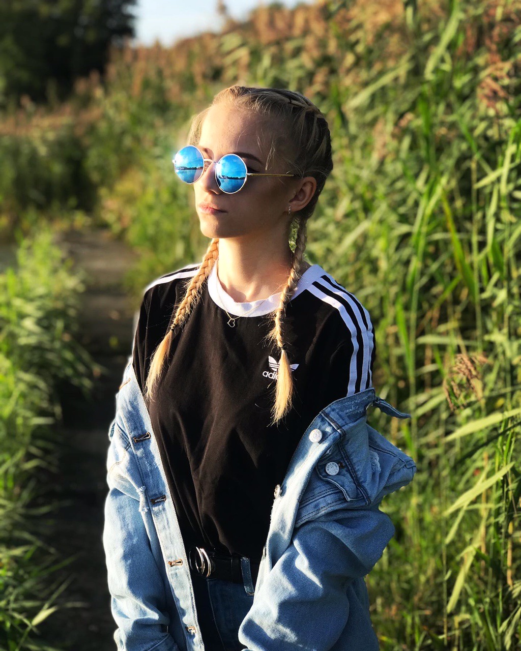 Portret młodej kobiety w okularach przeciwsłonecznych z odblaskami nieba i trzcin, ubrana w czarny t-shirt z białymi paskami i jeansową kurtkę, stojącej na tle wysokich traw.