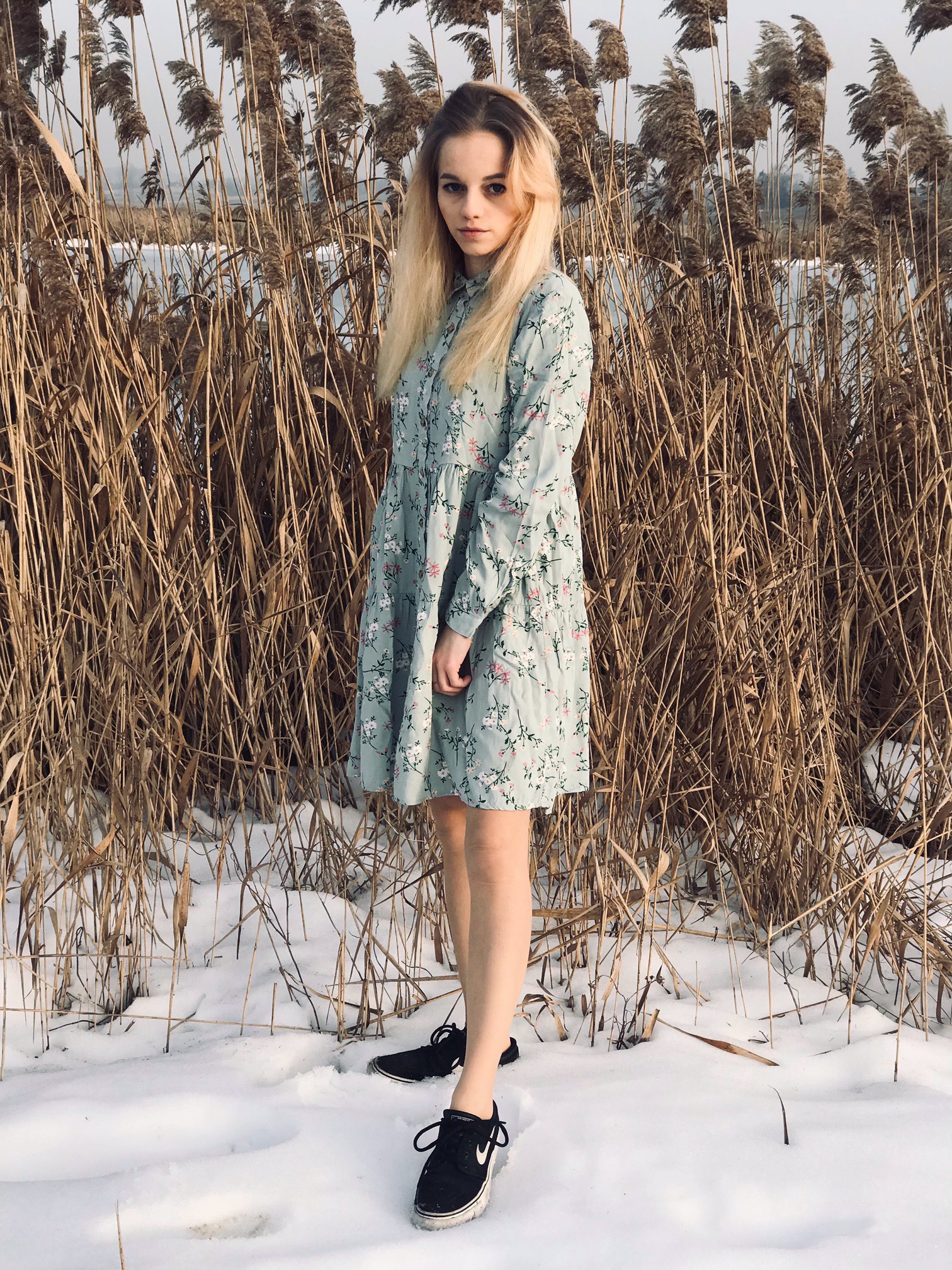 Portret młodej kobiety w kwiecistej sukience na tle suchych trzcin i śniegu, z lekkim uśmiechem i spojrzeniem w kamerę.