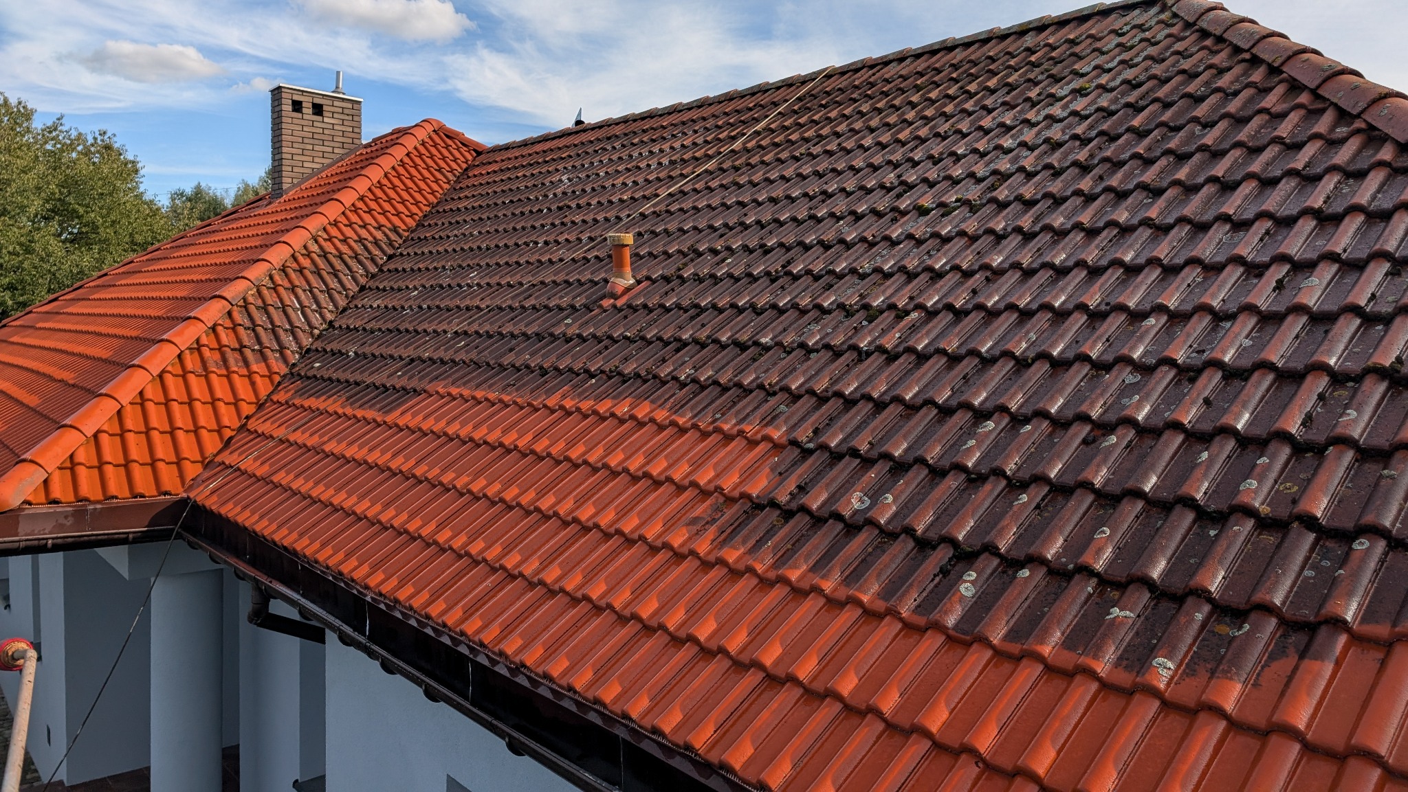 Częściowo wyczyszczony, czerwony dach z dachówki ceramicznej, widoczna wyraźna różnica w kolorze między brudną i czystą częścią, fragment komina w tle, lekko zachmurzone niebo.