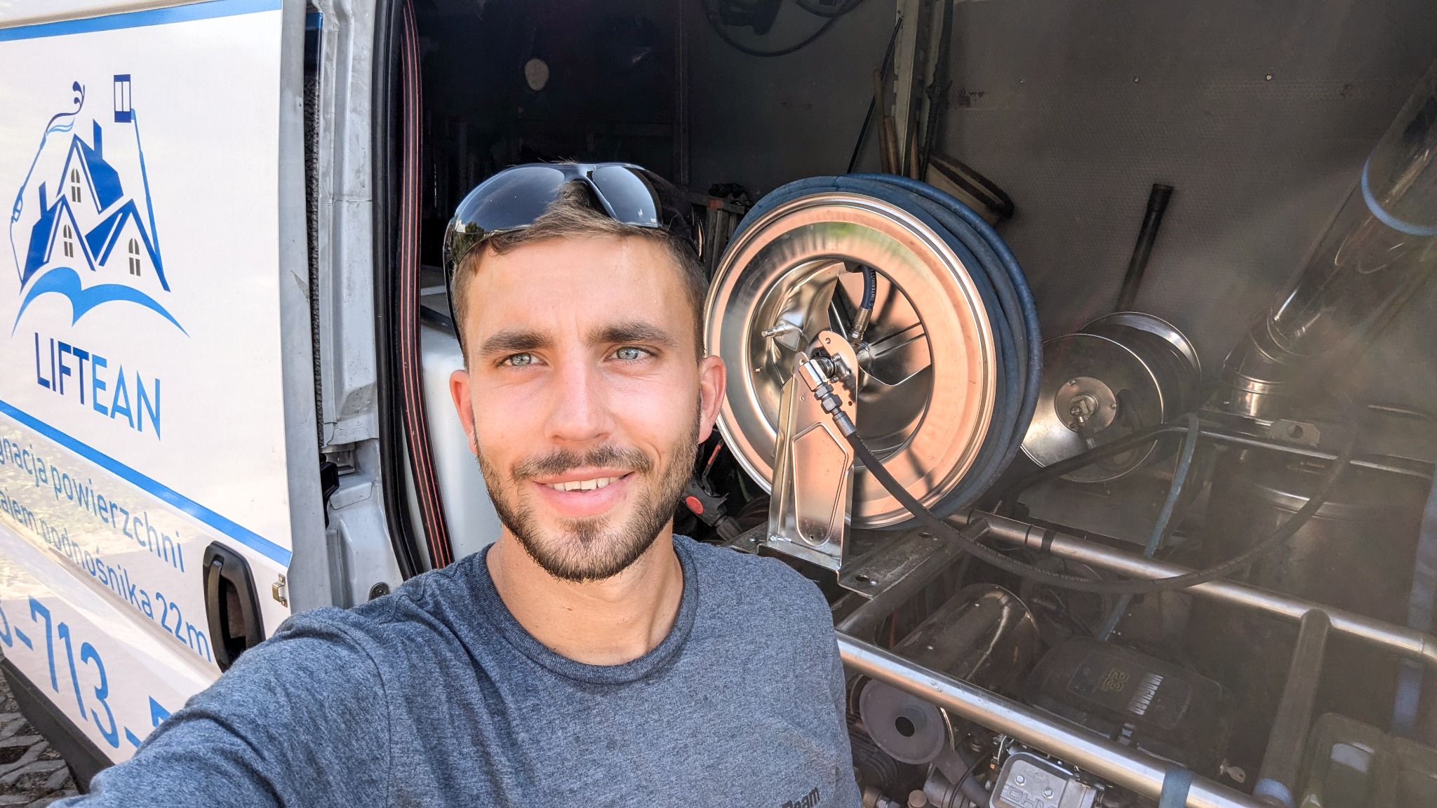 Uśmiechnięty mężczyzna w szarej koszulce i okularach na głowie, pozujący na tle wozu serwisowego z logiem firmy Liftean i sprzętem do czyszczenia powierzchni, w tym zwinięty wąż i turbina.