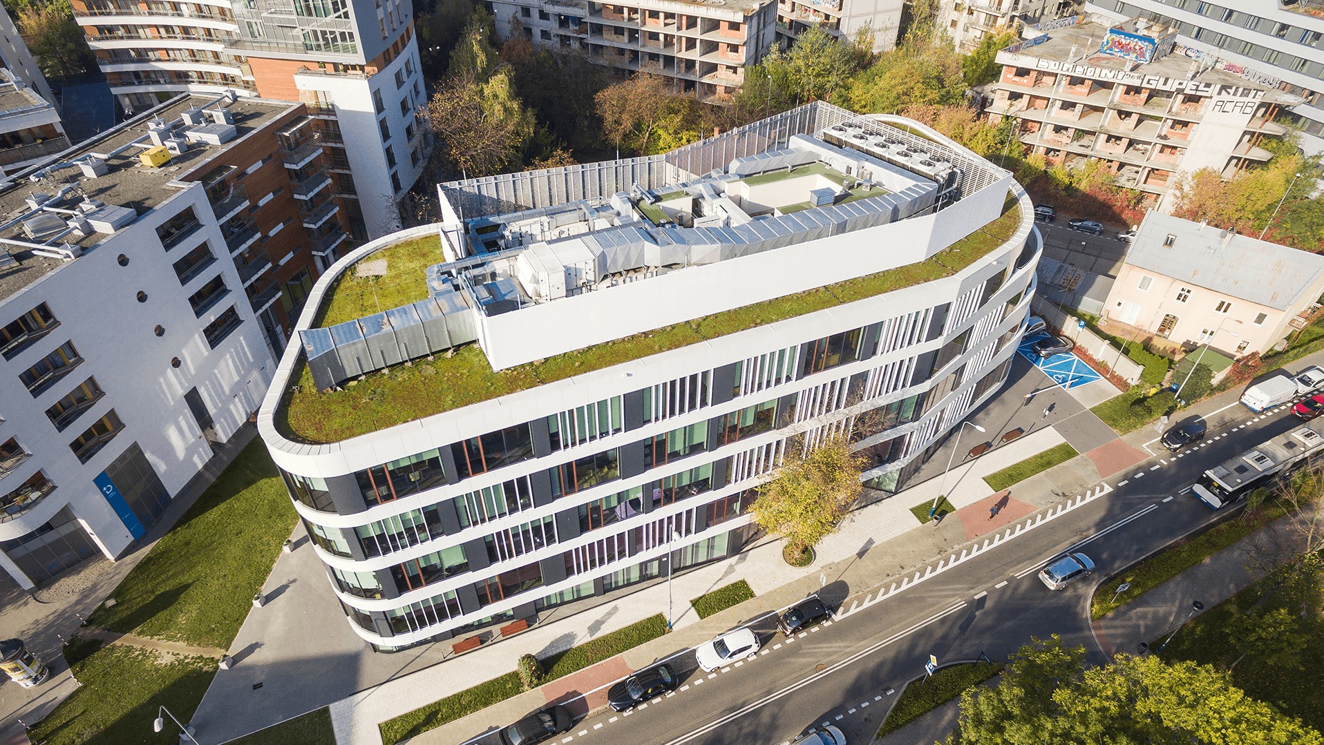 Widok z lotu ptaka na nowoczesny, biały budynek biurowy z zielonym dachem, systemami wentylacyjnymi na dachu, otoczony zielenią i ulicami z samochodami.