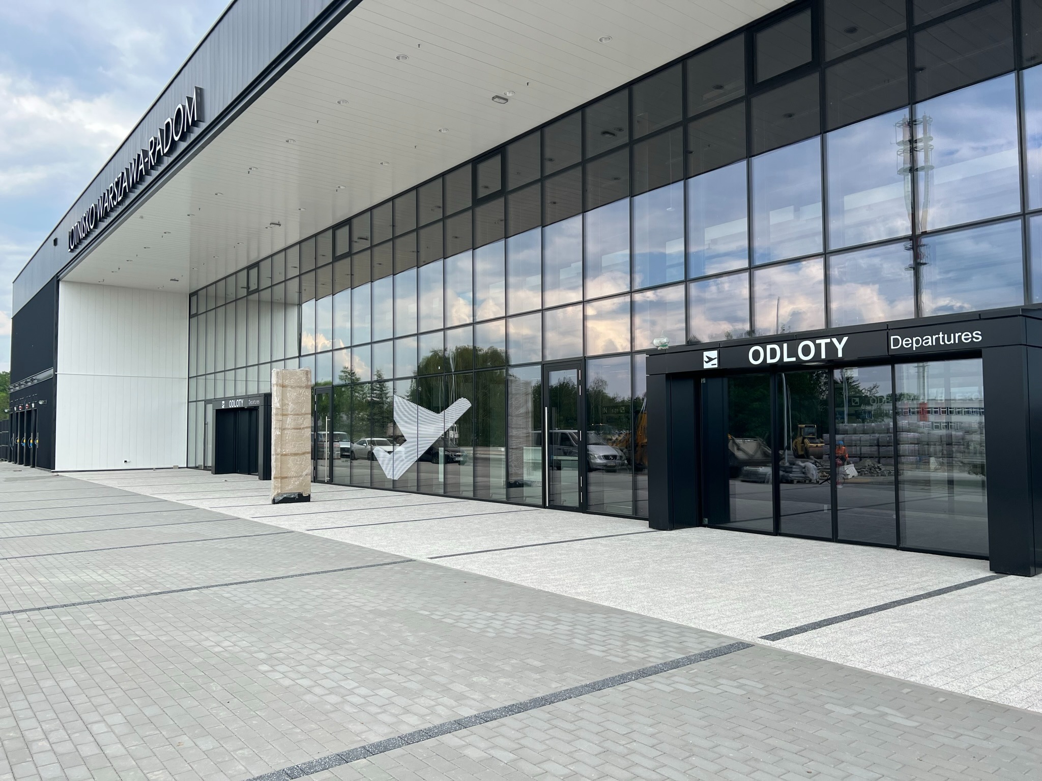 Nowoczesny budynek lotniska z dużymi szklanymi fasadami i napisem 'ODLOTY Departures' nad wejściem, widoczne prace budowlane w tle.
