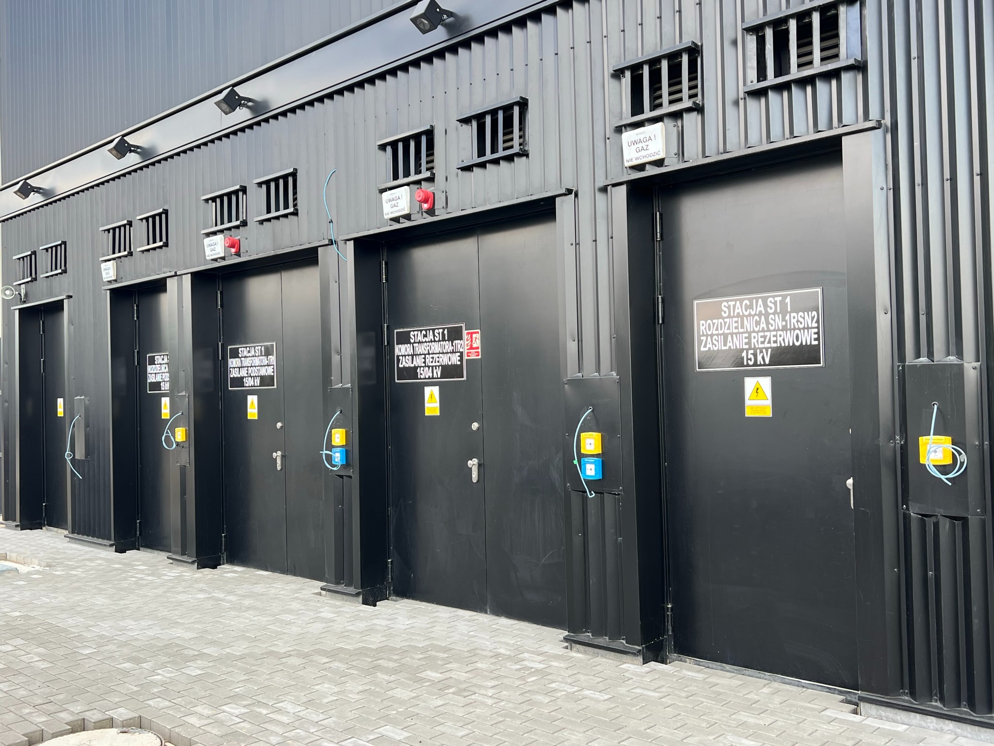 Zewnętrzny widok stacji transformatorowych w Krakowie, z czarnymi metalowymi drzwiami oznaczonymi tabliczkami informacyjnymi i ostrzegawczymi, oraz kratkami wentylacyjnymi u góry.
