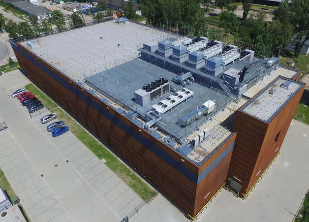 Widok z góry na dach budynku przemysłowego z zainstalowanymi systemami wentylacyjnymi i klimatyzacyjnymi, parking z samochodami w tle.