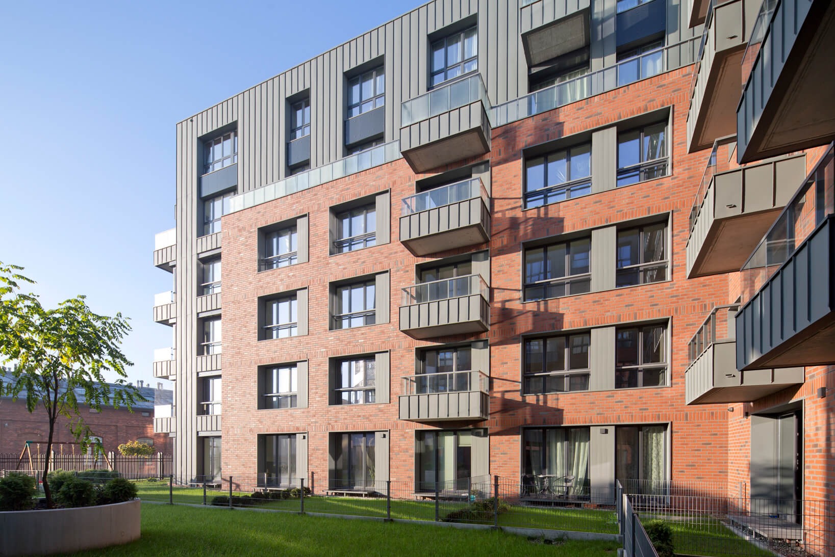 Nowoczesny budynek mieszkalny z ceglaną elewacją, metalowym wykończeniem i balkonami, otoczony zielonym trawnikiem i drzewami.