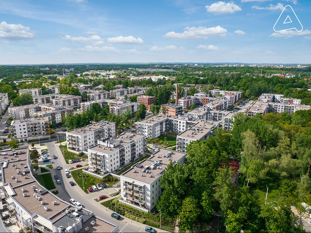 Osiedle nowych budynków wielorodzinnych z balkonami, widok z lotu ptaka w słoneczny dzień, zieleń drzew w tle.