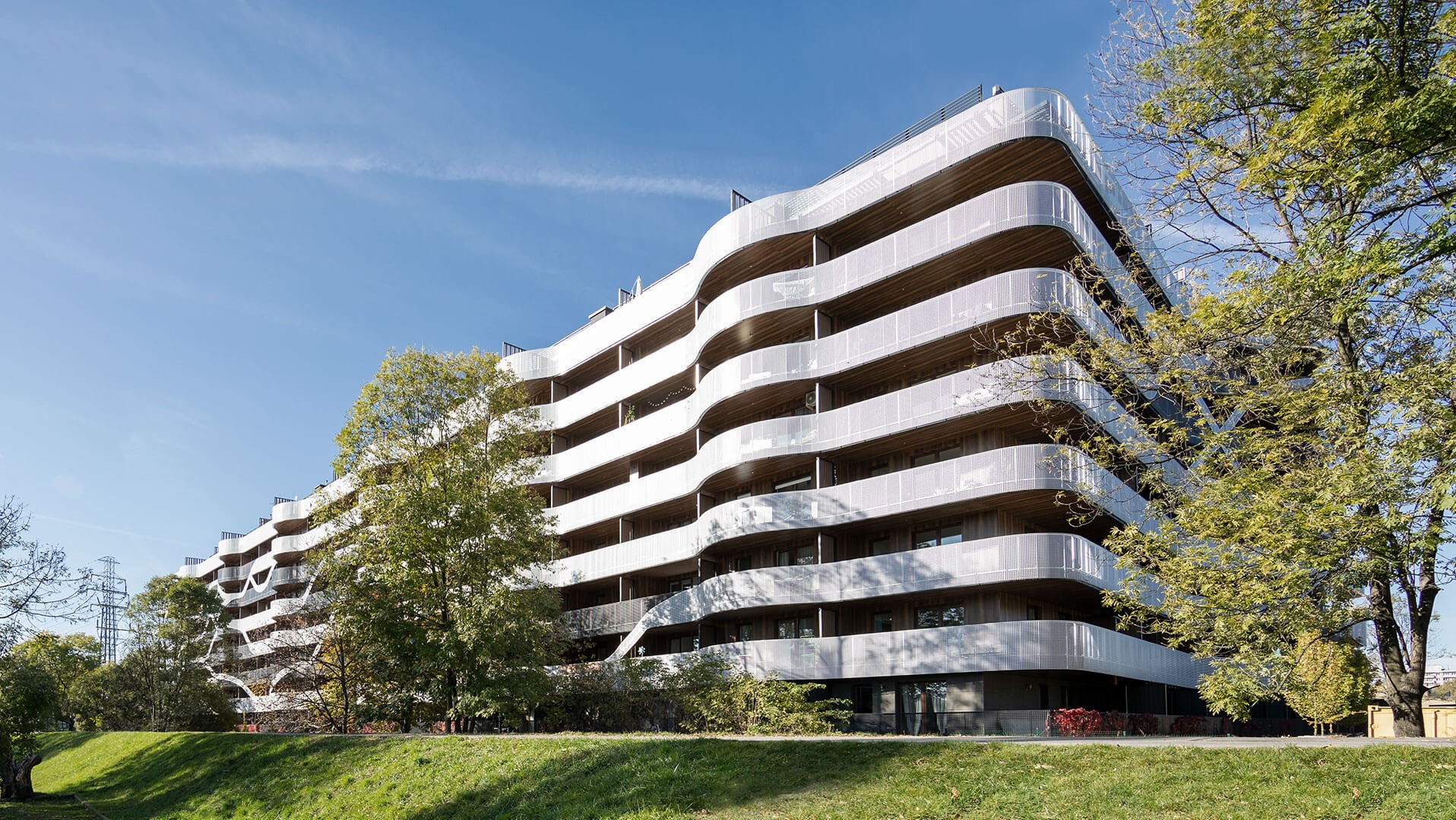 Wielopiętrowy budynek mieszkalny z falującymi balkonami i perforowaną elewacją, widoczny z perspektywy trawnika w słoneczny dzień.