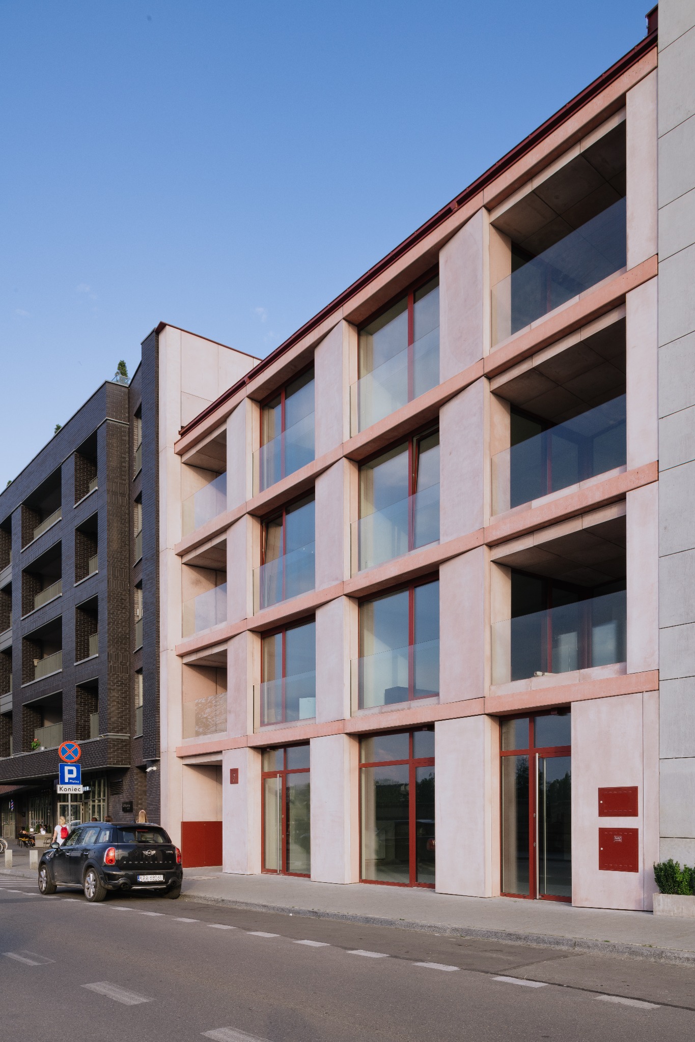 Nowoczesny budynek mieszkalny z balkonami, jasna elewacja z czerwonymi ramami okien i drzwiami, obok ciemniejszy budynek z cegły, zaparkowany samochód na ulicy.
