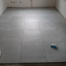 Układanie szarych płytek podłogowych w trakcie remontu kuchni, widoczna paczka z klejem i fragmenty niezabezpieczonej ściany.