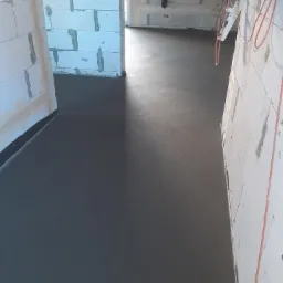 Świeżo wylana, gładka, ciemnoszara posadzka w surowym wnętrzu budynku z białymi ścianami z betonu komórkowego i wystającymi przewodami elektrycznymi.