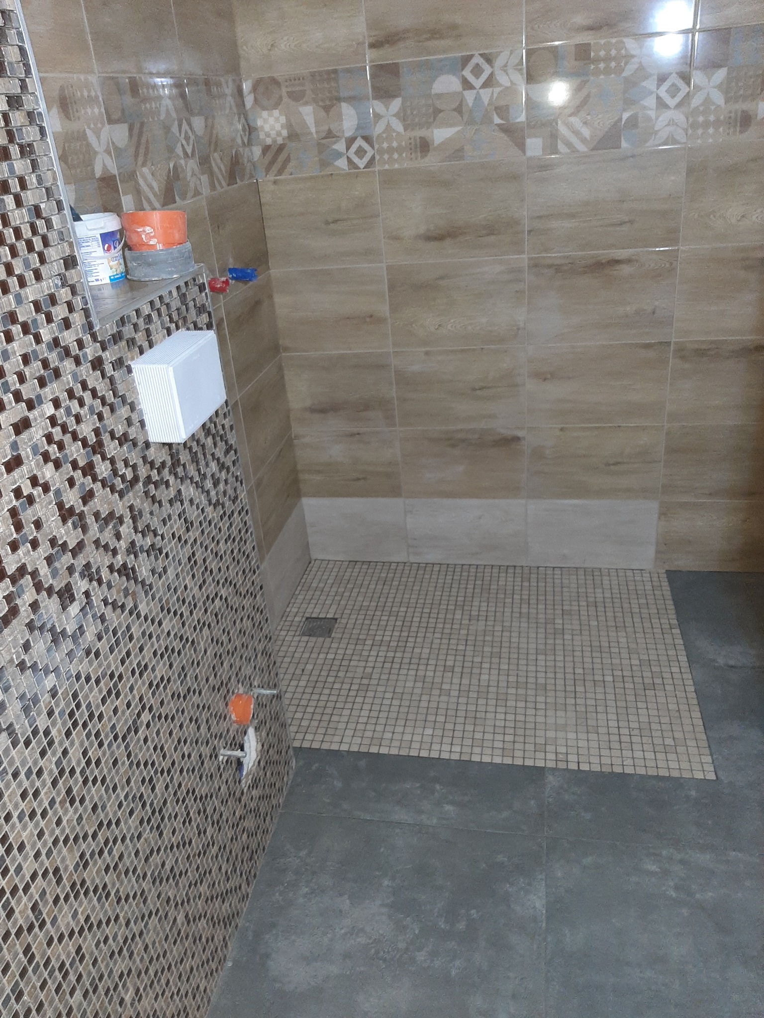 Wnętrze kabiny prysznicowej z mozaiką na ścianie, płytkami imitującymi drewno na górze i drobną mozaiką pod prysznicem, widoczne wiaderka z farbą i narzędzia glazurnicze.