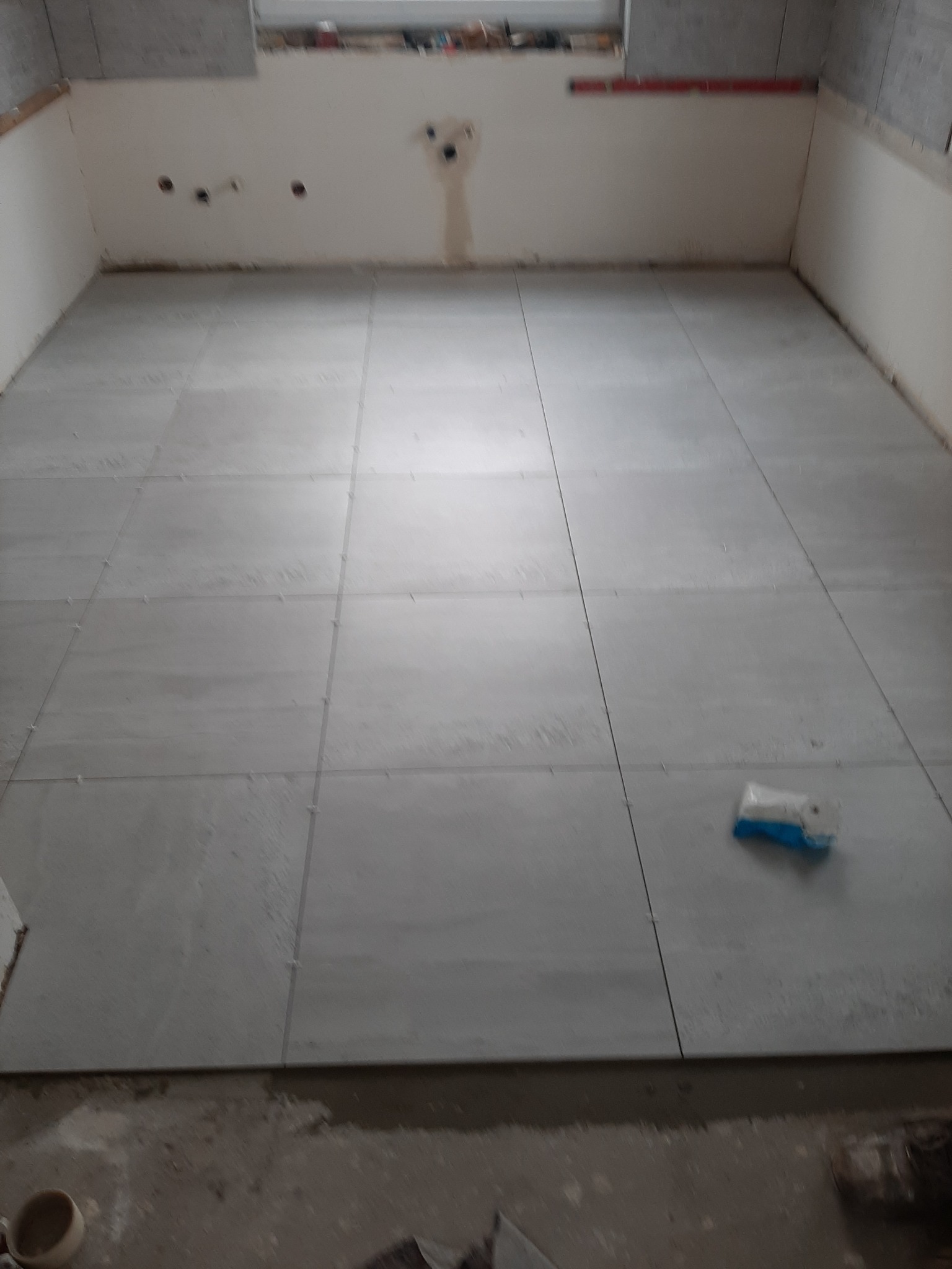 Układanie szarych płytek podłogowych w trakcie remontu kuchni, widoczna paczka z klejem i fragmenty niezabezpieczonej ściany.