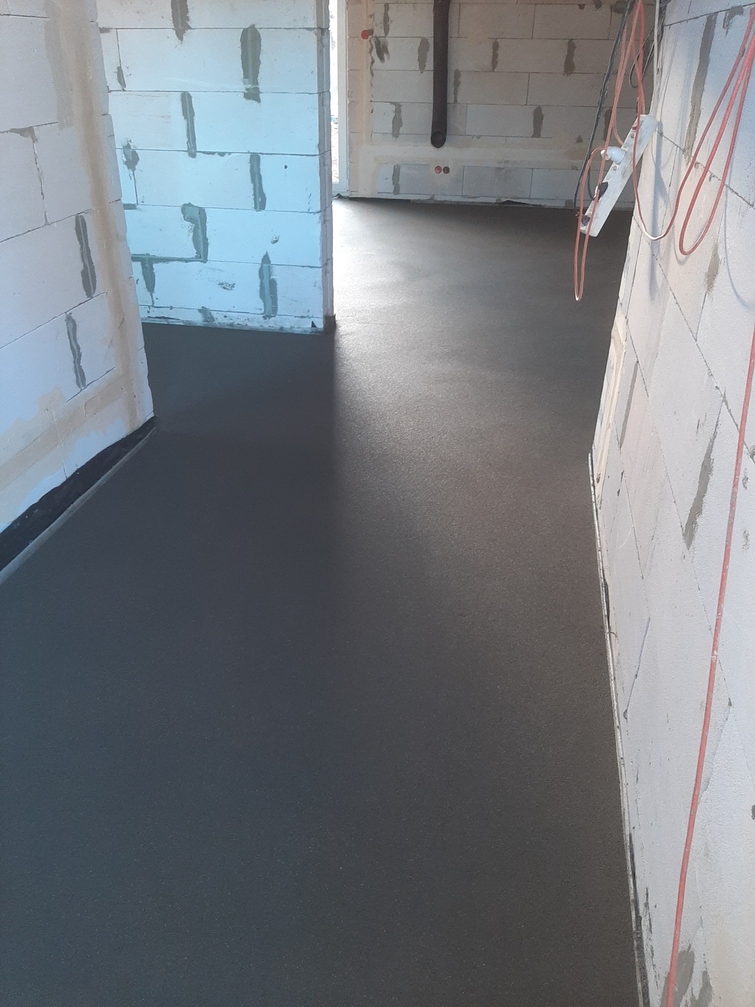 Świeżo wylana, gładka, ciemnoszara posadzka w surowym wnętrzu budynku z białymi ścianami z betonu komórkowego i wystającymi przewodami elektrycznymi.