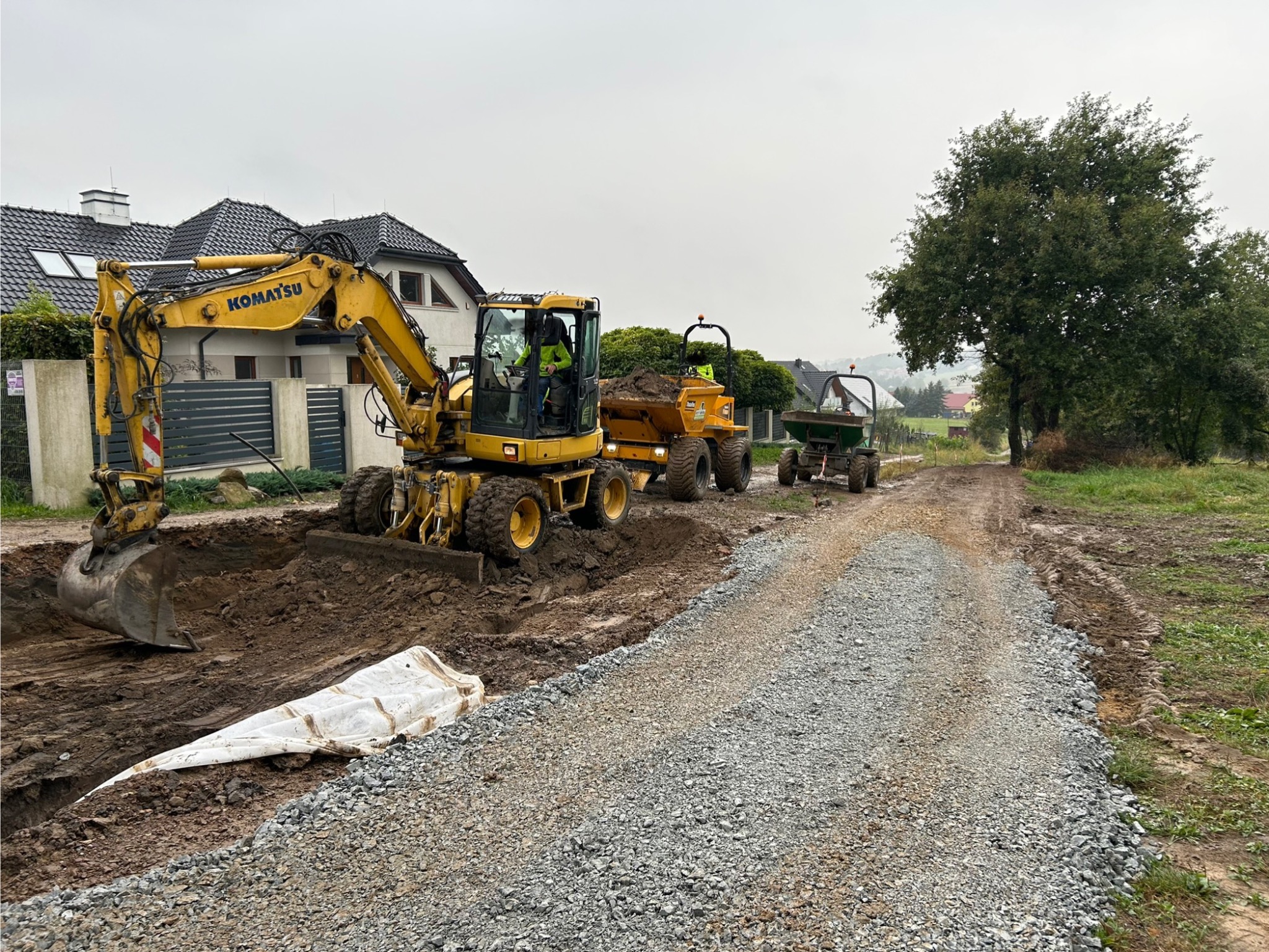 Żółta koparka Komatsu wykonuje wykop w ziemi, w tle dwie wywrotki, a w pierwszym planie utwardzona żwirowa droga