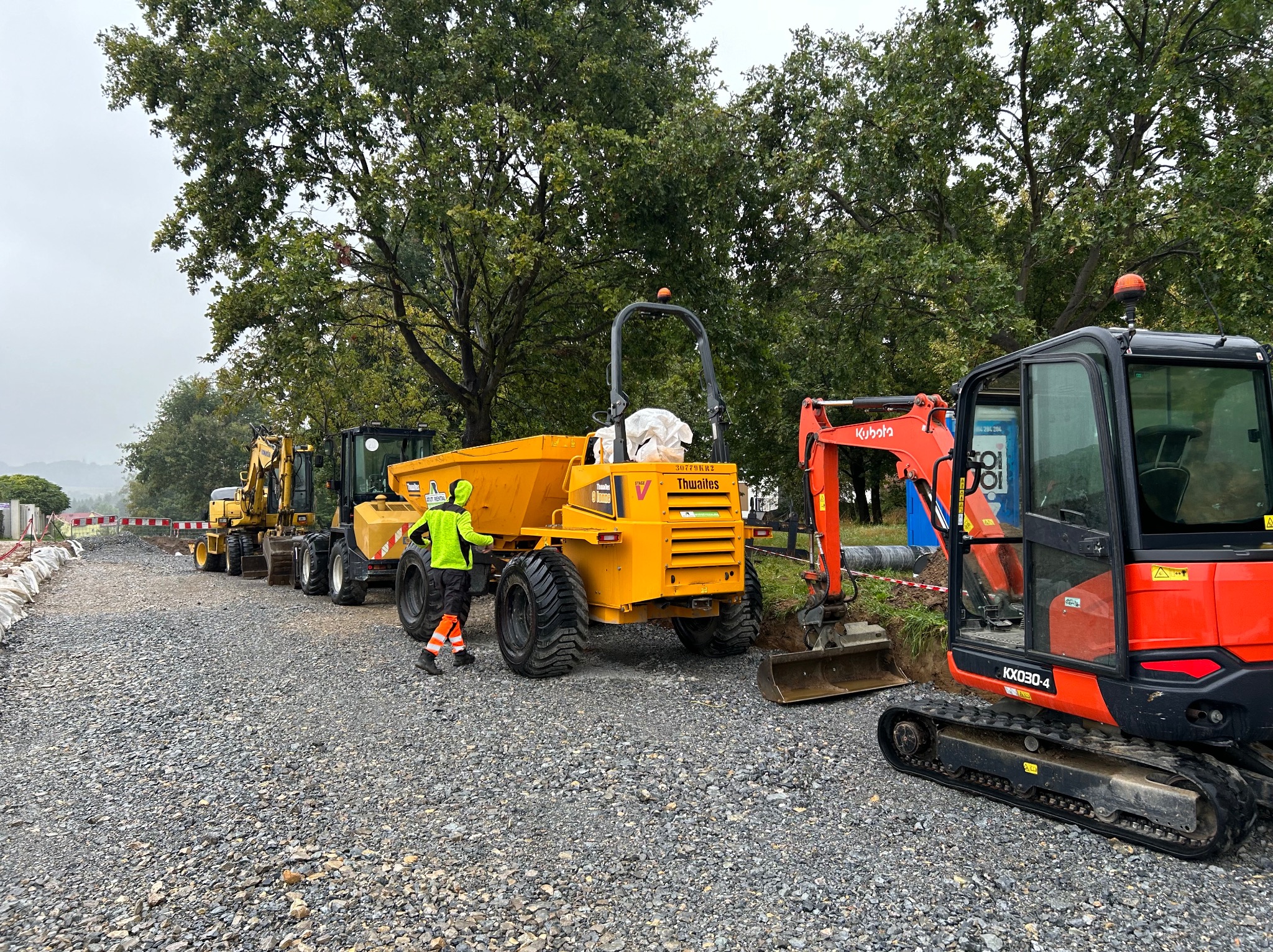Żwirowa droga w trakcie utwardzania z rzędem maszyn budowlanych: koparki, wywrotka Thwaites, pracownik w odblaskowej kamizelce i pomarańczowych nogawkach nadzorujący prace.