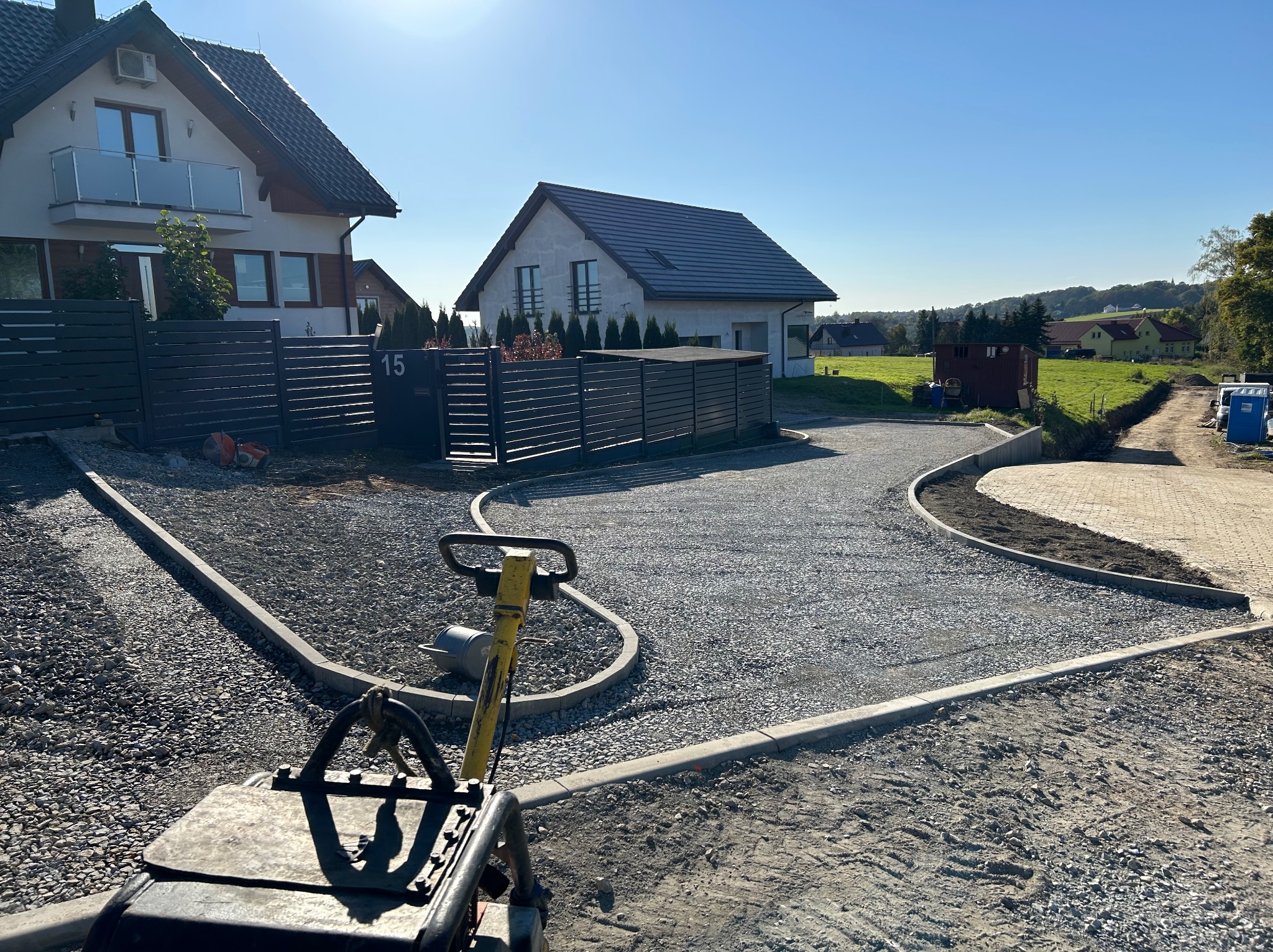 Przygotowanie podłoża pod kostkę brukową na posesji: ubijarka wibracyjna na żwirowym podłożu, obrzeża betonowe, w tle domy jednorodzinne i błękitne niebo.