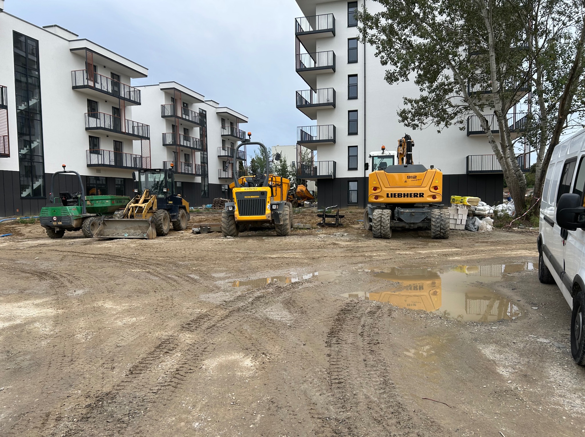 Żółte maszyny budowlane na placu budowy, w tle białe budynki z balkonami, teren błotnisty z kałużami. Widoczne ślady opon.